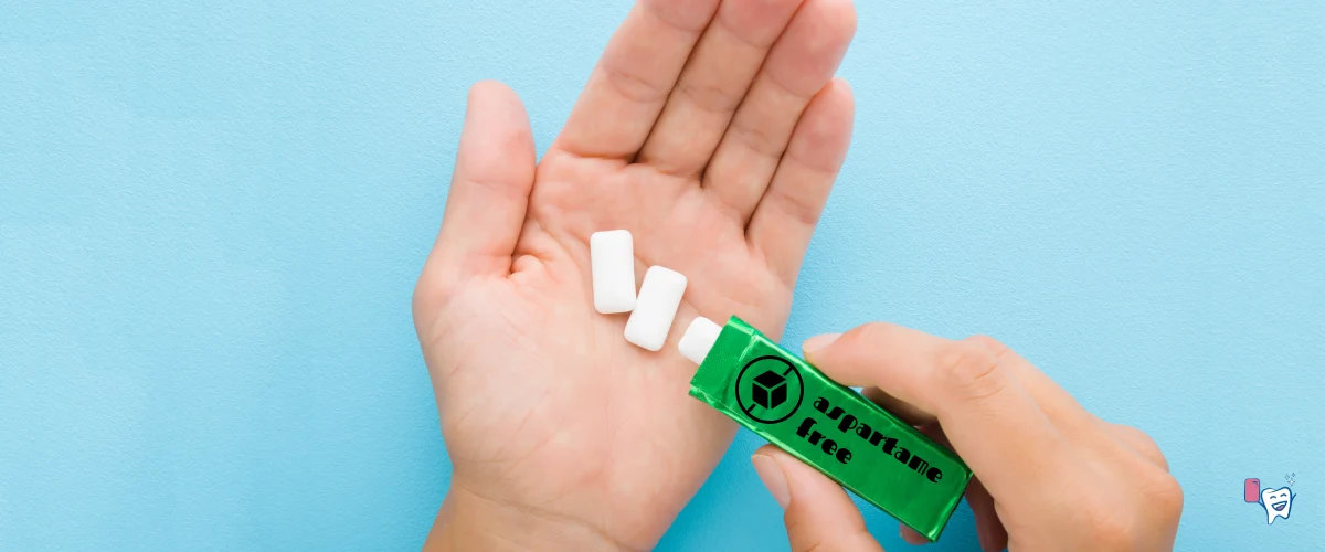 Picture of an open left hand holding white dragee chewing gum spilling out of a green pack with a black symbol and the description aspartame free in the right hand against a light blue background | For the article - Aspartame free chewing gum | web: HealthyChewingGum.com | source: shutterstock | Edited by: Jiri Samuel