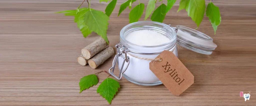 Abgebildet ist Xylit-Zucker (Xylitol) in einem durchsichtigen Glasgefäß mit einem Textetikett auf braunem Papier "Xylitol" mit einem Hintergrund auf einem Holztisch mit Birkenblättern und Birkenholzstücken | Für Artikel: Xylitol | Für die Website: healthychewinggum.com | Quelle: shutterstock