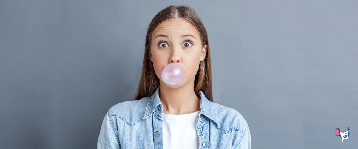 The picture shows a girl blowing bubble with chewing gum, wearing a blue jacket with white T-shirt under it, standing in front of a grey background | For article: Chewing Gum for Slimmer Face | For website: healthychewinggum.com | Source: shutterstock