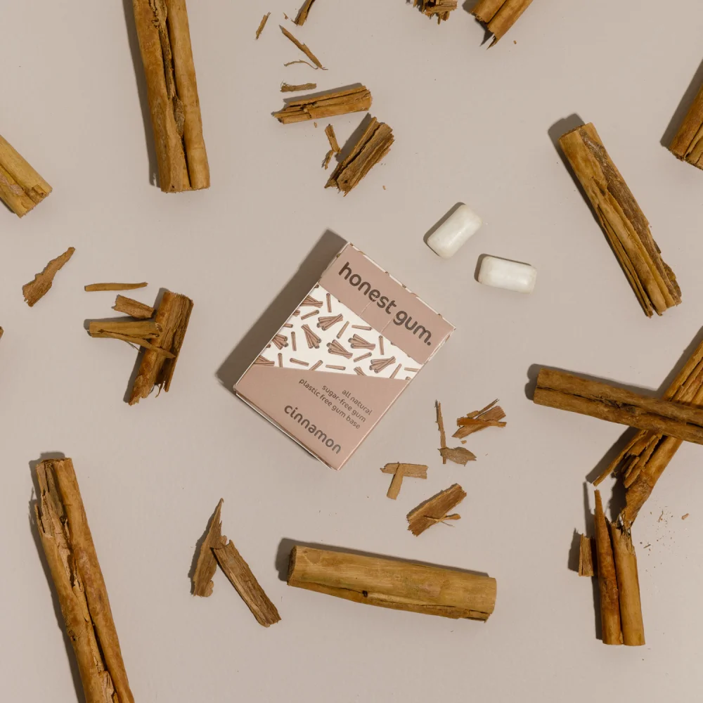obrázek žvýkačky skořicové přírodní žvýkačky od honest gum ve krabičce s 12 ks zepředu na pozadí s naaranžovanou skořicí a žvýkačkami | Žvýkačky bez plastu, bez cukru, udržitelné a bez aspartamu | Pro web: healthychewinggum.com | Zdroj: honest gum