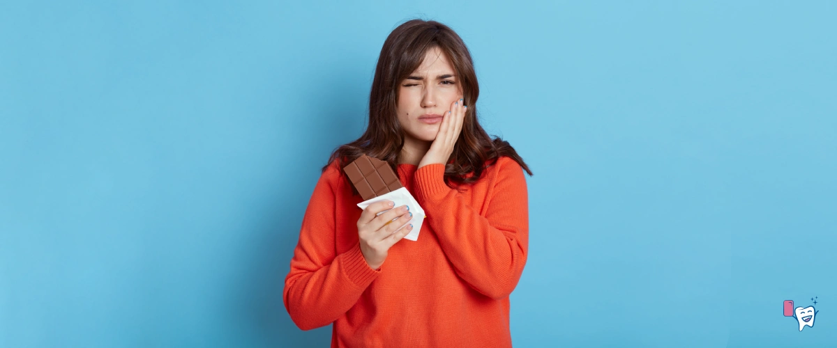 The picture shows a girl in orange hoodie having toothache and holding a chocolate bar in one hand, standing in front of a blue background | For article: How Sweets lead to Tooth Decay | For website: healthychewinggum.com | Source: shutterstock