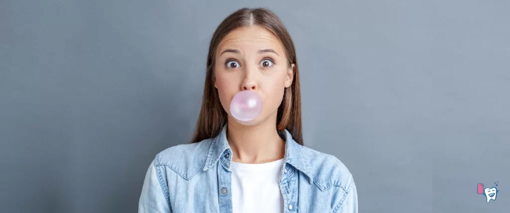 Na obrázku je dívka, která fouká bubliny se žvýkačkou, na sobě má modrou bundu a pod ní bílé tričko a stojí před šedým pozadím | Pro článek: Jak zhubnout v obličeji | Pro webové stránky: healthychewinggum.com | Zdroj: shutterstock