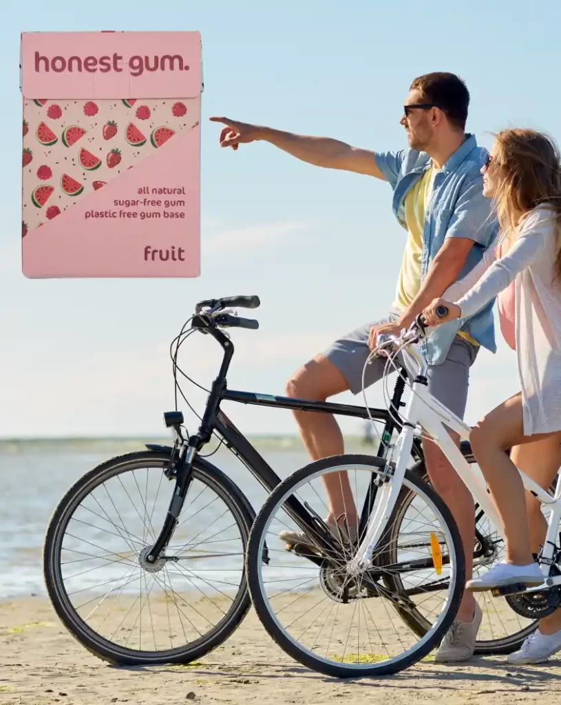 Junges Paar sitzt auf Fahrrädern und fährt an einem schönen sonnigen Tag am Strand in der Nähe des Meeres | Banner mit 100 % natürlichem, ehrlichem Kaugummi | Für die Website: healthychewinggum.com | Autor: Jiri Samuel | Hintergrundfotoquelle: Shutterstock.com