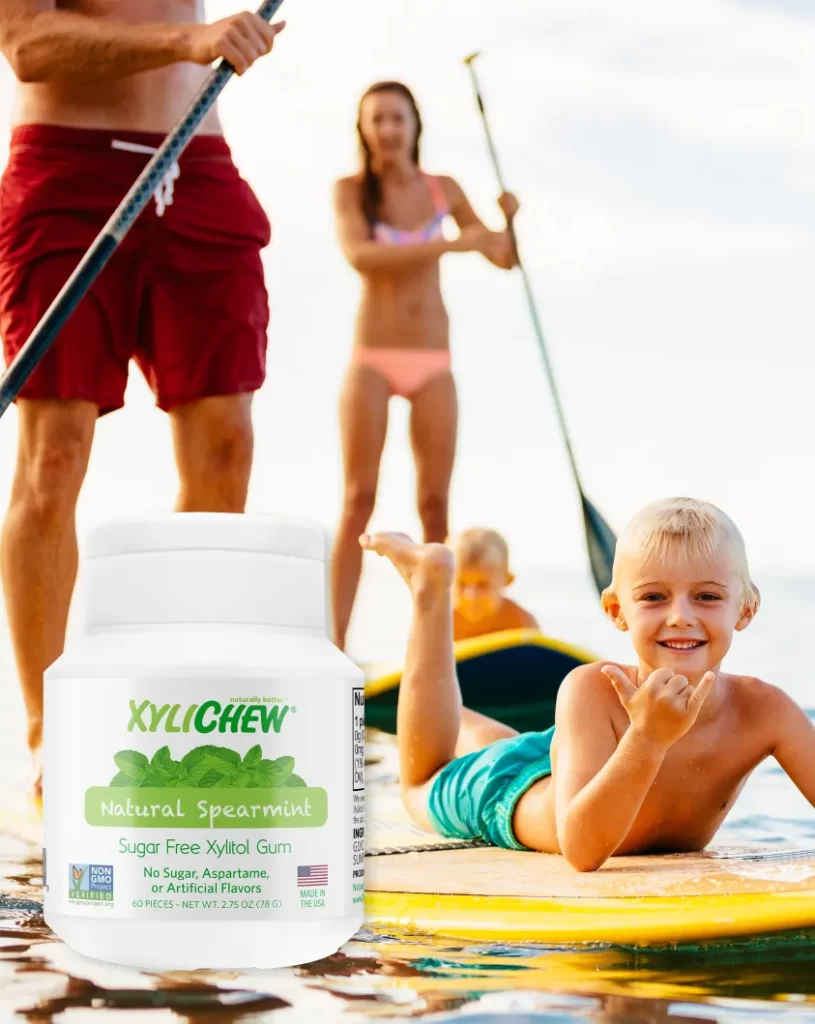 Glückliches Kind auf einem Paddleboard im Meer an einem sonnigen Tag mit seinem Bruder und seinen Eltern, mit Mama und Papa im Hintergrund | Banner mit Xylichew – Grüne Minze | Für die Website: healthychewinggum.com | Autor: Jiri Samuel