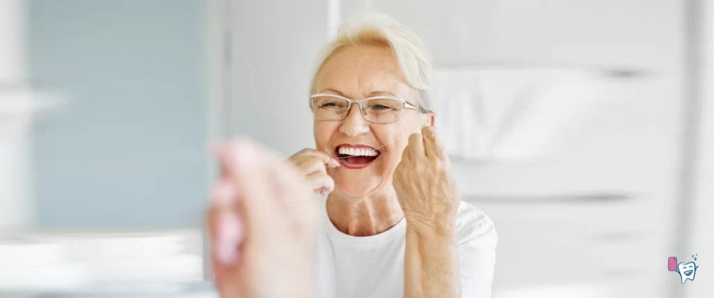 An elderly woman with glasses is happily flossing her healthy white teeth by looking in a mirror | For article: Why Oral Hygiene Matters? | For website: healthychewinggum.com | Source: shutterstock