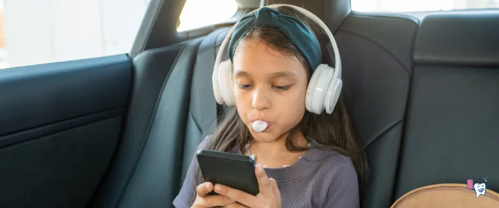 The image shows a school-going girl sitting on the back seat of a car. She is chewing a gum while she is engrossed in her phone | For article: Positive Effects of Frequent Gum Chewing | For website: healthychewinggum.com | Source: shutterstock