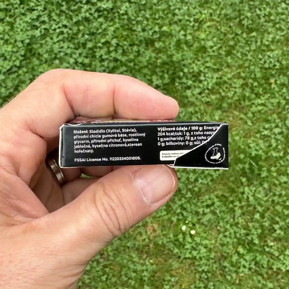Aktivní uhlí Spearmint (mátové) přírodní žvýkačky Gud Gum bez plastových materiálů a bez škodlivé chemie | Držené v ruce levou rukou na pozadí trávník zahrady - pohled z boku na krabičku žvýkaček - viditelný text složení a výživové údaje | Bez aspartamu, bez cukru, bez sorbitolu, bez BHT, bez BHA | Žvýkačky pro děti