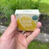 Wrapper of Lemon Chewing Gum in front with activated charcoal, gum in palm with activated charcoal in back held in left hand with mint, grass and dirt in garden in background.