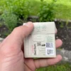 Lemon gum Gud Gum in a box from the back held in the left hand with herbs, grass and a garden bed in the background