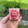 Wrapper of Raspberry Chewing Gum in front with activated charcoal, gum in palm with activated charcoal in back held in left hand with mint, grass and dirt in garden in background.