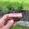 Wrapper of Raspberry Chewing Gum in front with activated charcoal, gum in palm with activated charcoal in back held in left hand with mint, grass and dirt in garden in background.