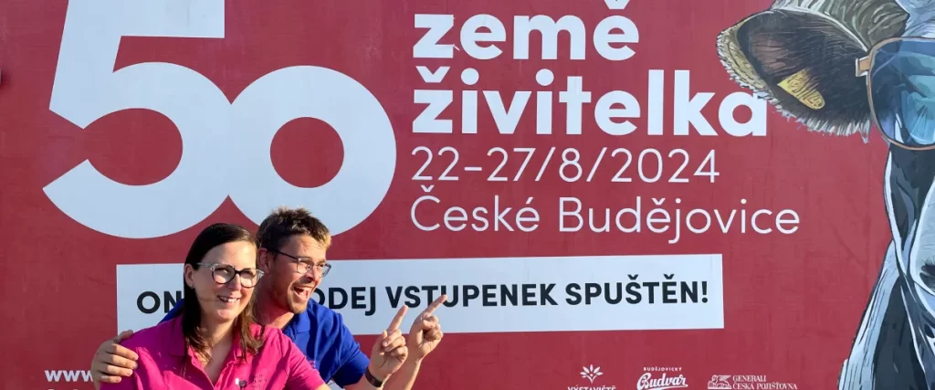 Fotografie mladé ženy a mladého muže stojící před bilboardem výstavy Země živitelka 2024 v Českých Budějovicích ukazujíc levou rukou na online prodej vstupenek - že je již spuštěn usmívajíc se a těšíc se na tuto výstavu kde budou mezi vystavovateli - kde budeme prodávat zdravé žvýkačky - těšíme se na vaši návštěvu | Pro: Pozvánka na výstavu Země živitelka 2024 na výstaviště České Budějovice | Autor fotografie a úprav: Jiří Samuel | Pro web: healthychewinggum.com