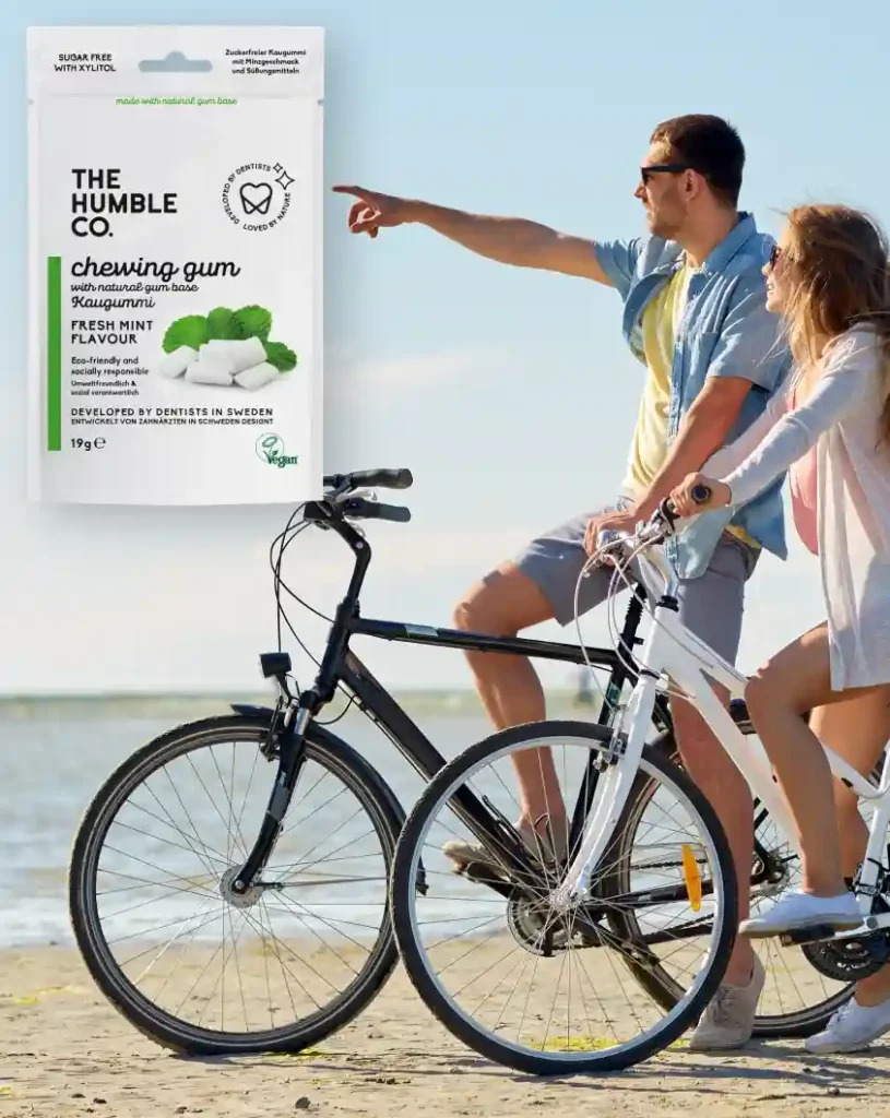 Mladý pár sedí na bicykloch a jazdí po pláži pri mori v krásny slnečný deň | Banner so 100 % prírodnou zdravou žuvačkou od spoločnosti The Humble Co. | Pre webovú stránku: healthychewinggum.com | Autor: Jiri Samuel | Zdroj fotografie v pozadí: shutterstock.com