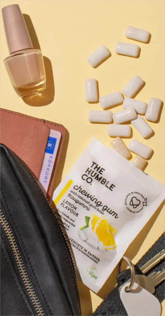 Spilled healthy gum from a bag by Swedish brand The Humble Co. lying next to other items from a woman's purse with nail polish and a wallet containing her ID card | Photo presenting Swedish gum brand The Humble Co. with lemon flavor | Source: The Humble Co. | For the website: healthychewinggum.com