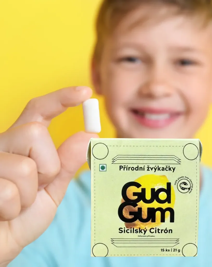 A young, smiling boy wearing a light blue T-shirt, holding white pieces of gum on the finger of his right hand, with a yellow screen print in the lower right corner in the background—Gud Gum children's gum